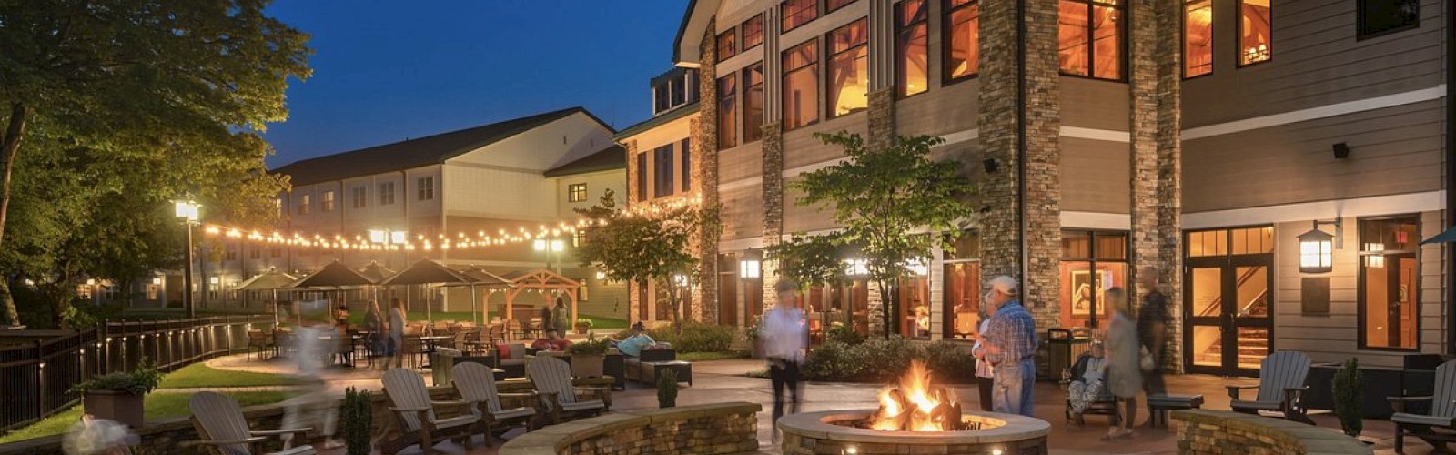 An outdoor lounge area at night with a fire pit, string lights, and seating. People are relaxing near a large building under a clear sky.