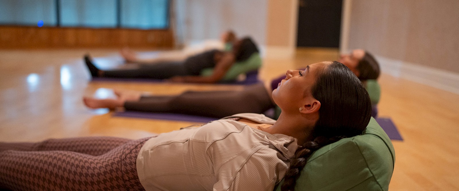 People lying on yoga mats in a peaceful room, possibly meditating or resting, with focus on a woman in the foreground.