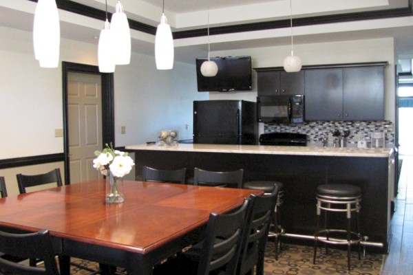 A modern kitchen and dining area with a wood table, chairs, pendant lights, a black and white kitchen with bar stools, and a wall-mounted TV.