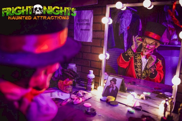 The image shows a person in a clown costume and makeup sitting at a vanity mirror with lights, part of Fright Nights Haunted Attractions.