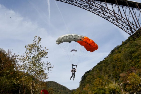 A person is parachuting near a large bridge, surrounded by trees and mountains under a clear sky.