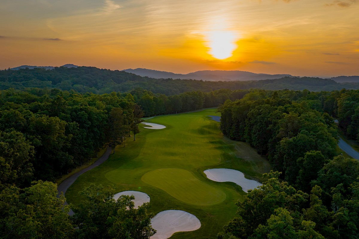 A golf course with sand traps and a lush fairway surrounded by trees at sunset, casting a warm glow over the landscape.
