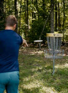 A person wearing a blue shirt is playing disc golf in a forest, aiming at a metal disc golf basket.