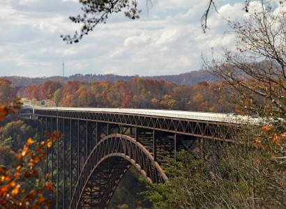 This is an image of a large steel arch bridge spanning a forested valley with autumn foliage, under a partly cloudy sky.