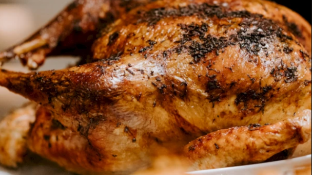 The image shows a roasted chicken with crispy, golden-brown skin, seasoned with herbs, served on a white plate.