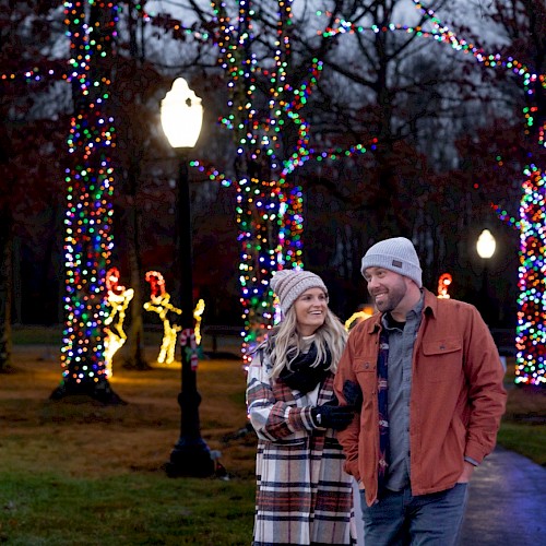 A couple walks through a park decorated with colorful holiday lights on trees and glowing figures, creating a festive atmosphere.