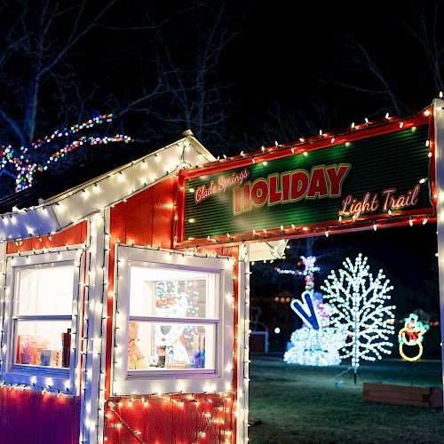 A festive holiday light display featuring a small booth, illuminated trees, and colorful decorations, creating a cheerful atmosphere.