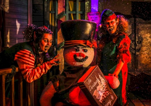 People dressed as elves pose with a snowman holding a Christmas-themed box in a festive setting.