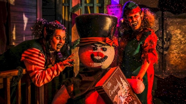 People dressed as elves pose with a snowman holding a Christmas-themed box in a festive setting.