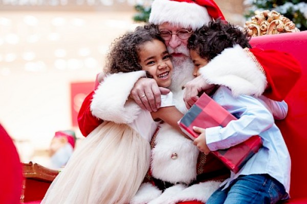 Santa Claus hugging two happy children, one holding a gift, with festive decorations and a Christmas tree in the background.