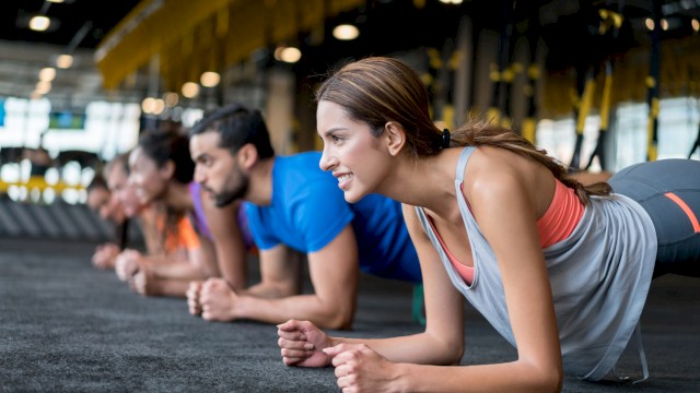 A group of people in a gym performing planks on a dark mat. They are aligned in a row, focused, and wearing athletic clothing.