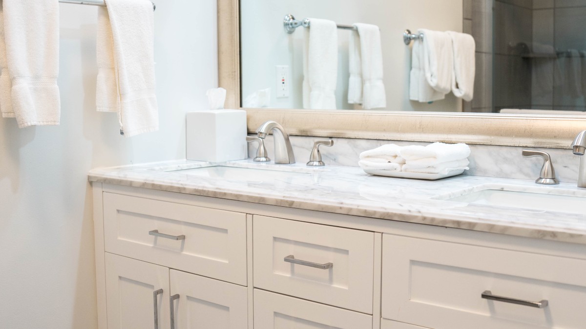 A modern bathroom with a large mirror, white vanity, and neatly folded towels on a rack. A bath mat is placed on the dark tile floor.