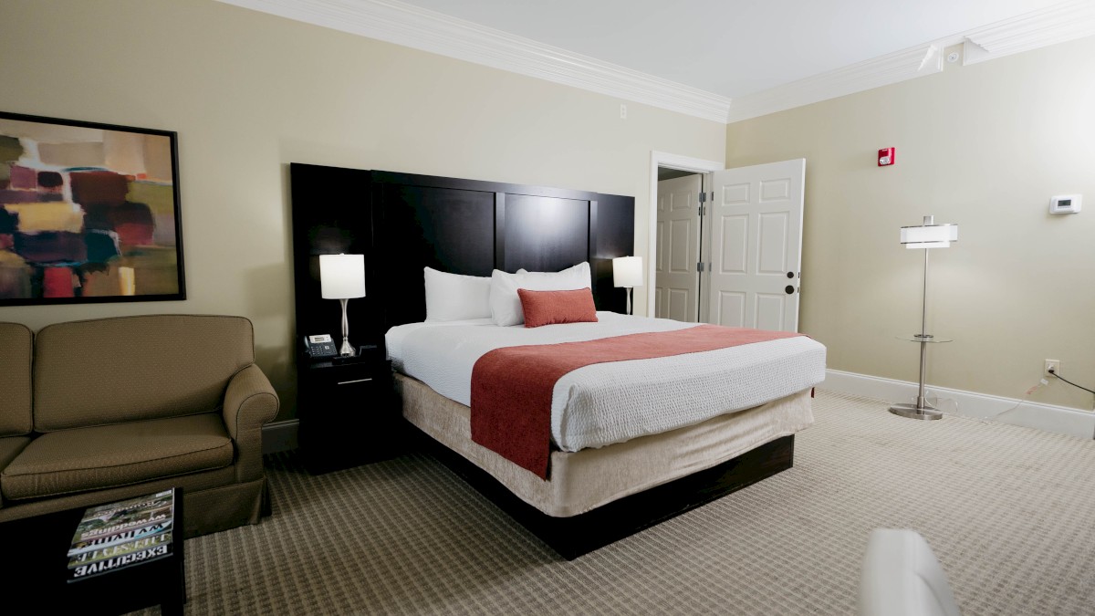 A neatly made hotel room with a large bed, nightstands, lamps, a beige sofa, abstract art, and a standing lamp in a bright, clean space.