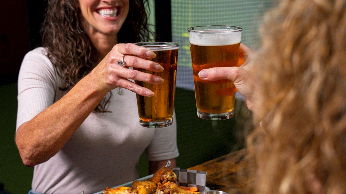 A group of people toast with beers while enjoying sandwiches and chips at a table. Screens in the background display digital graphics.