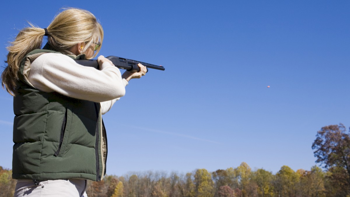 A person is outdoors aiming a shotgun, wearing a green vest and aiming at a distant target against a clear blue sky.