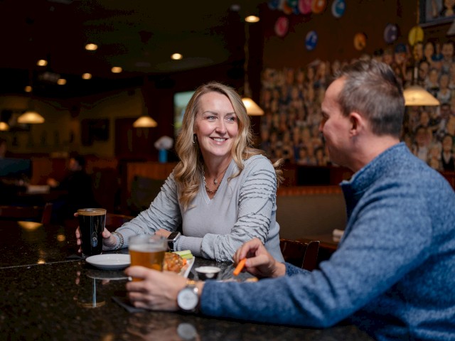 A woman and a man are sitting at a bar, enjoying drinks and a meal. The setting is warm and well-lit, with wall art in the background.