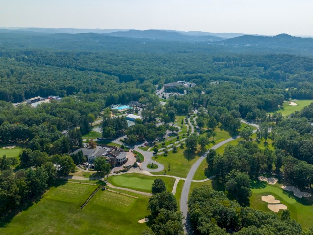 An aerial view of a lush landscape featuring a golf course, roads, trees, and buildings set amidst rolling hills and expansive greenery.