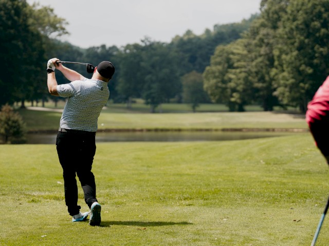 A golfer swings a club on a lush green course, with trees in the background, while another person watches nearby.
