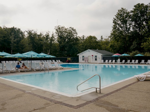 A rectangular outdoor pool with lounge chairs, umbrellas, and a small building, surrounded by trees and a cloudy sky.