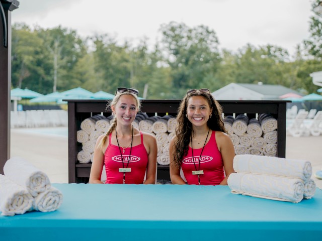 Two people in uniform sit at a table with rolled towels, likely by a poolside, with outdoor seating and greenery in the background.