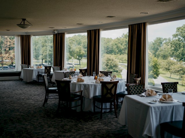 The image shows a dining area with tables set for a meal, featuring large windows with a view of a green landscape.
