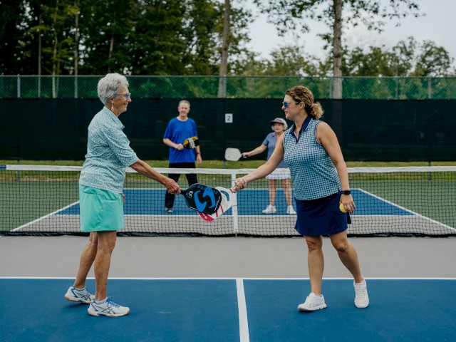 People playing pickleball, two shaking hands across the net. Others in the background hold paddles on an outdoor court.