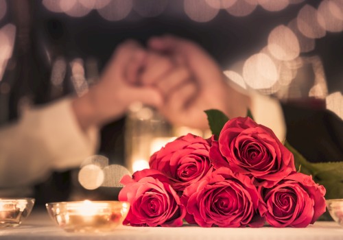 A bouquet of red roses on a dinner table with candles, blurred hands resting in the background, creating a romantic ambiance.