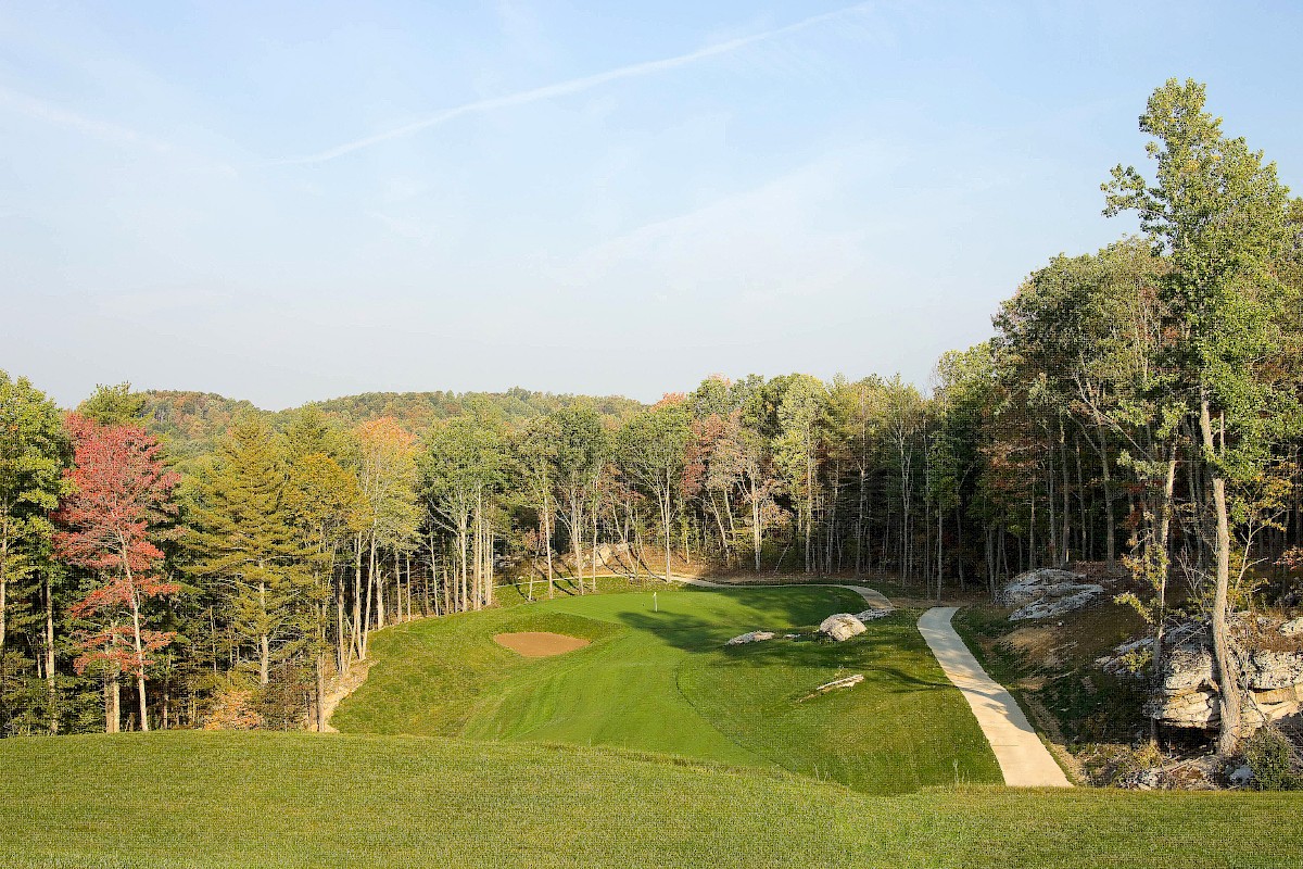 A scenic golf course set among trees with a green, fairway, bunkers, and a stone path on the right, under a clear blue sky.