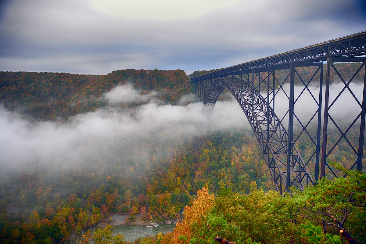 A steel arch bridge spans over a misty valley with lush autumn foliage, creating a scenic and atmospheric landscape view.