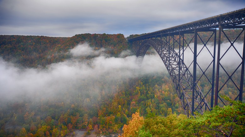 A steel arch bridge spans over a misty valley with lush autumn foliage, creating a scenic and atmospheric landscape view.