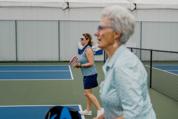 Two people are playing pickleball on an outdoor court. One is in the foreground with sunglasses, while the other is in the background.
