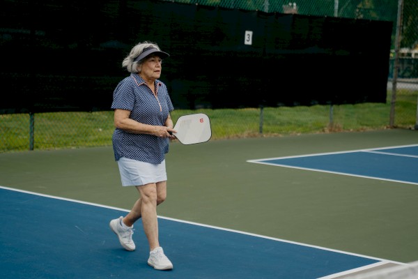 An older woman plays pickleball on an outdoor court, holding a paddle and wearing a visor, blue shirt, white shorts, and sneakers.