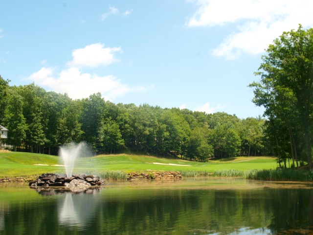 A serene landscape featuring a pond with a water fountain, surrounded by lush green trees under a partly cloudy sky.
