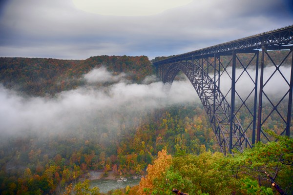 A large arch bridge spans over a foggy, forested valley with autumn foliage, creating a dramatic and scenic landscape.