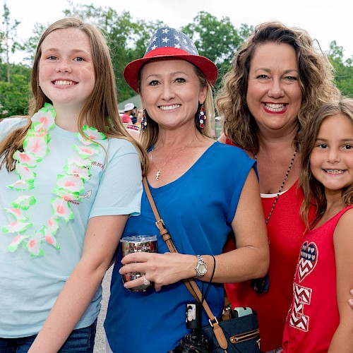 Four people are smiling at an outdoor event, with two adults and two children in casual attire.