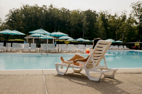 A person is relaxing on a lounge chair by a pool with umbrellas and trees in the background, creating a serene atmosphere.