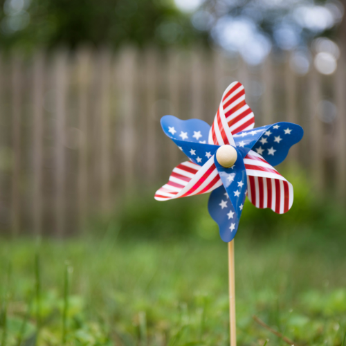 A pinwheel with a red, white, and blue star pattern stands in the grass, with a wooden fence blurred in the background.