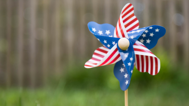 A pinwheel with a red, white, and blue star pattern stands in the grass, with a wooden fence blurred in the background.
