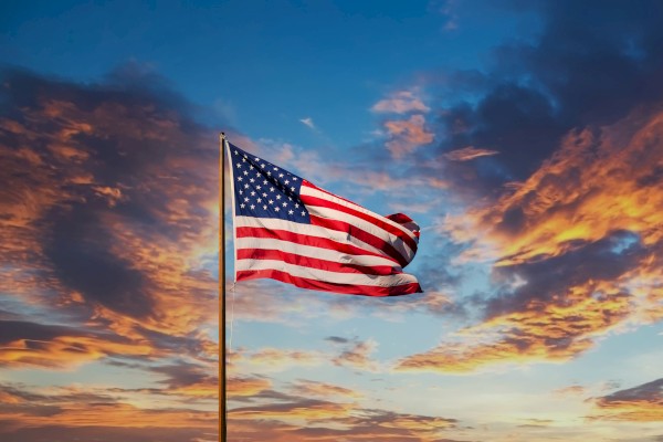 An American flag waves on a pole against a backdrop of a colorful, cloud-filled sky at sunset.