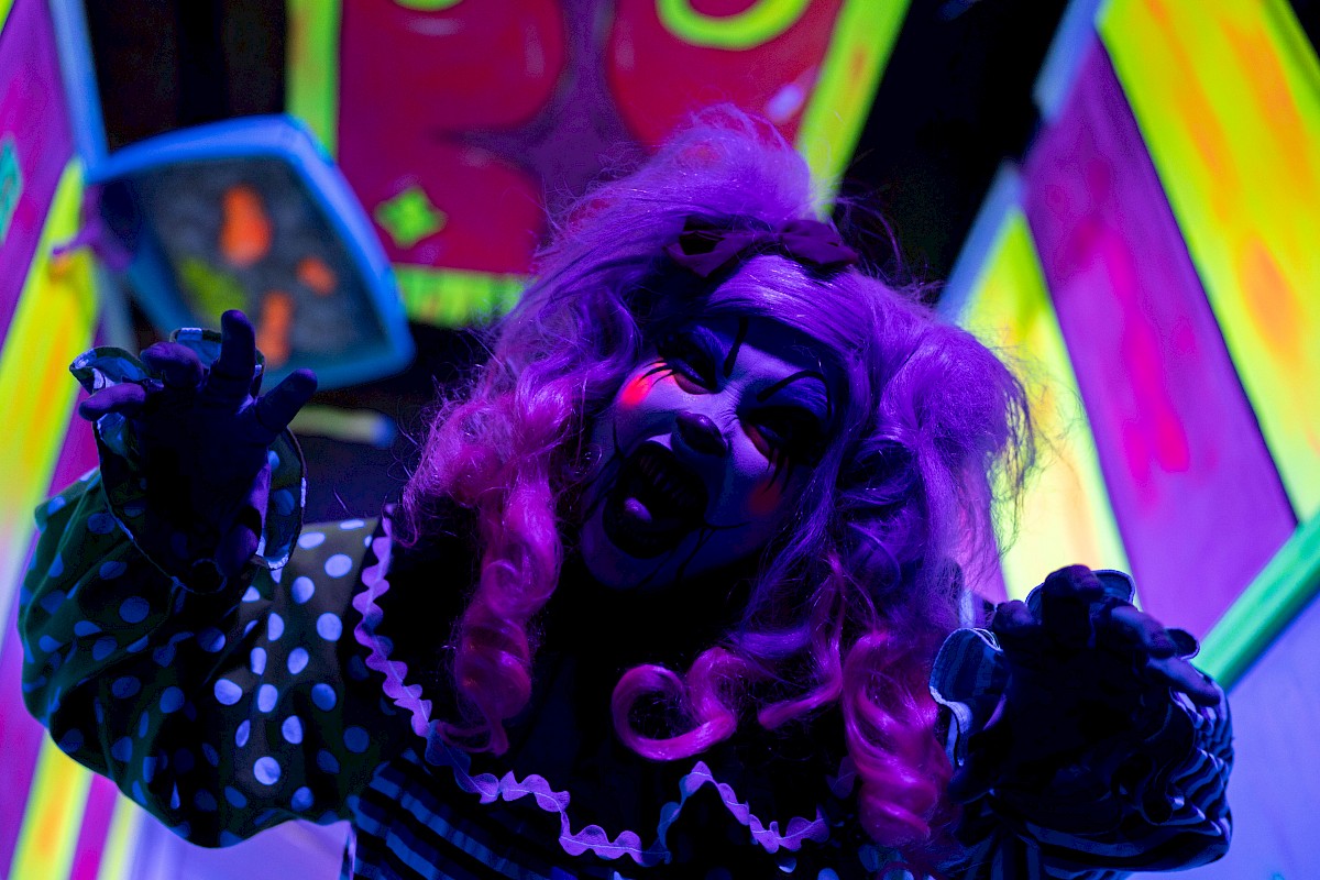 A person in spooky clown makeup and costume is in a neon-lit environment, posing dramatically with hands reaching out.