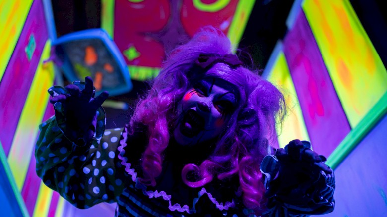 A person in spooky clown makeup and costume is in a neon-lit environment, posing dramatically with hands reaching out.