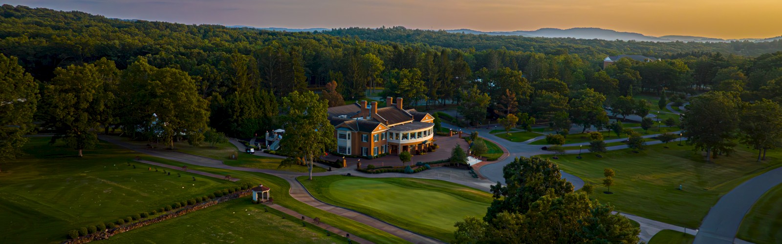 A scenic view of a golf course and clubhouse during sunset, surrounded by lush trees and rolling hills in the distance.
