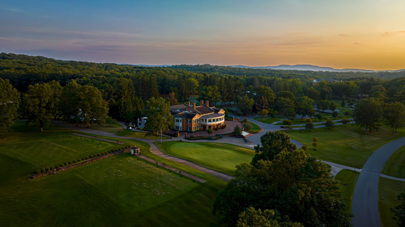 A scenic view of a golf course and clubhouse during sunset, surrounded by lush trees and rolling hills in the distance.