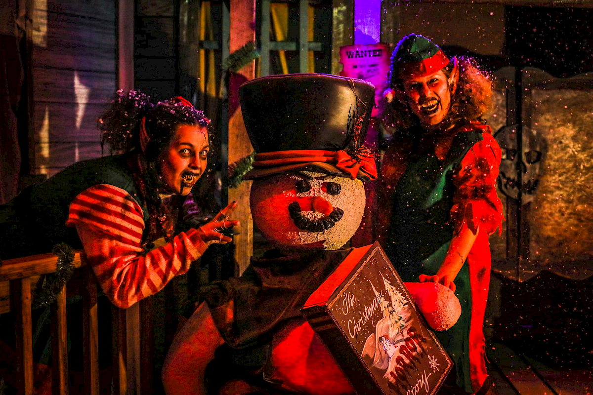 Two costumed kids and a neon-lit clown snowman carnival prop, with festive hats and a quirky box, in a dark, snowy scene. ending with a period.