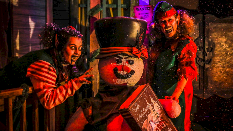 Two costumed kids and a neon-lit clown snowman carnival prop, with festive hats and a quirky box, in a dark, snowy scene. ending with a period.