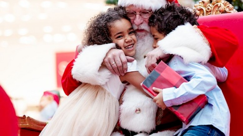 Kids hug Santa, joyful Christmas moment, wrapped gifts nearby, festive lights and trees in the background.