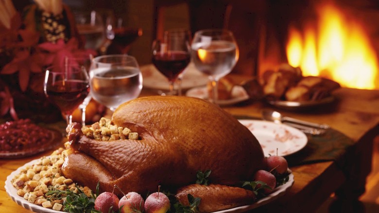 A festive dinner table features a roasted turkey centerpiece with vegetables, wine glasses, and a glowing fireplace in the background.