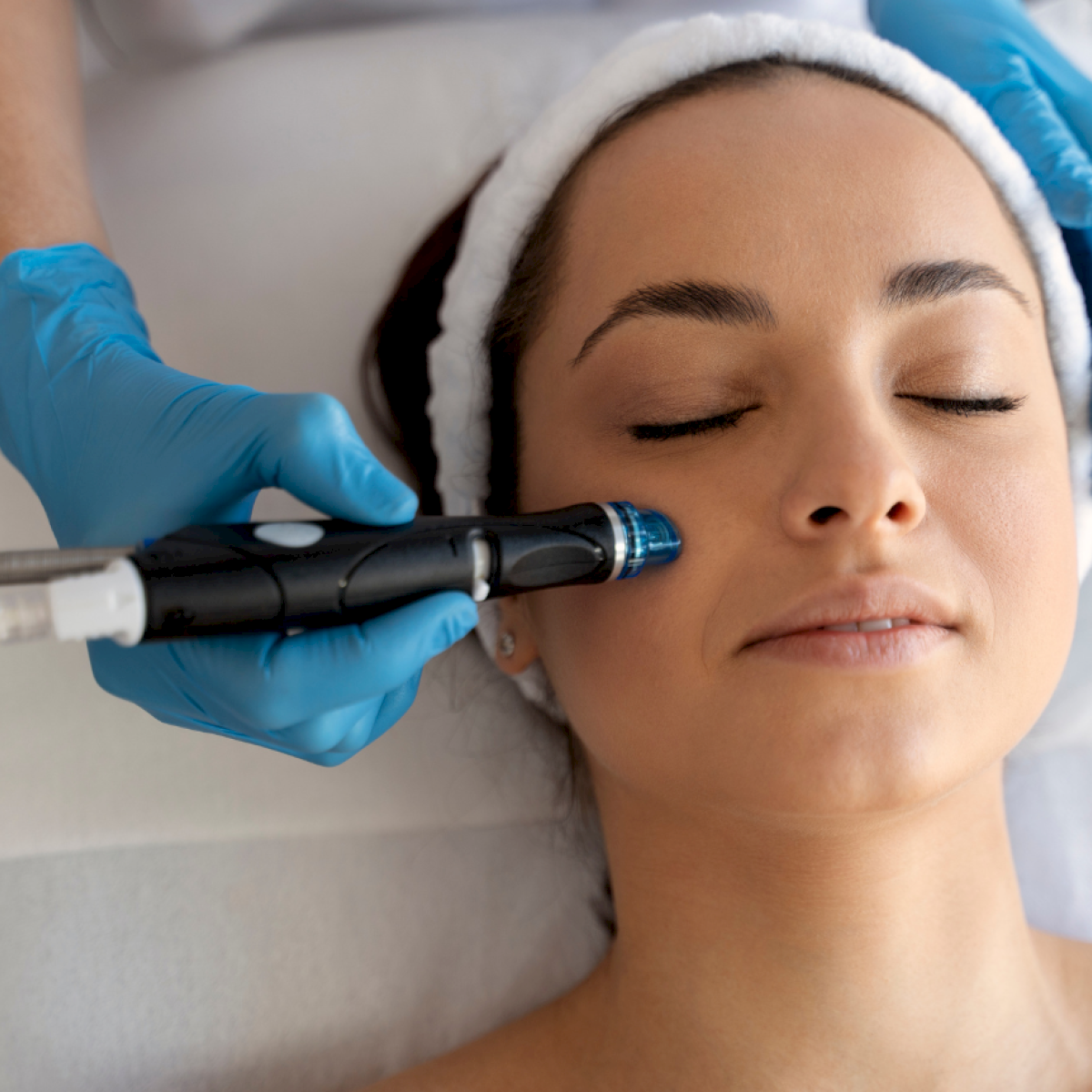 A woman lies with eyes closed as a practitioner uses a handheld device on her cheek, wearing blue gloves and a headband during a cosmetic facial treatment.