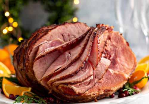 A sliced roasted ham centerpiece on a plate, garnished with orange slices and herbs, festive background lights, flavorful and juicy.