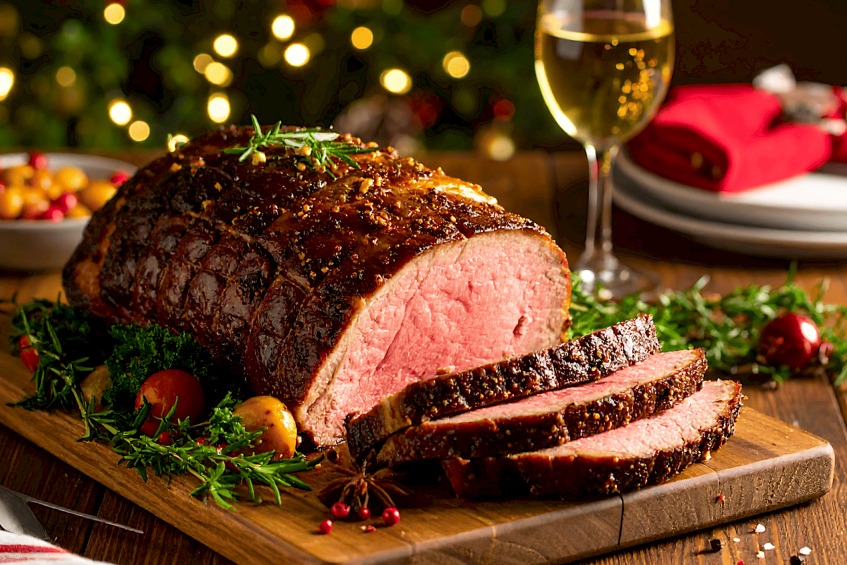A juicy glazed beef roast on a cutting board with sliced portions, herbs, and a festive setting with wine.